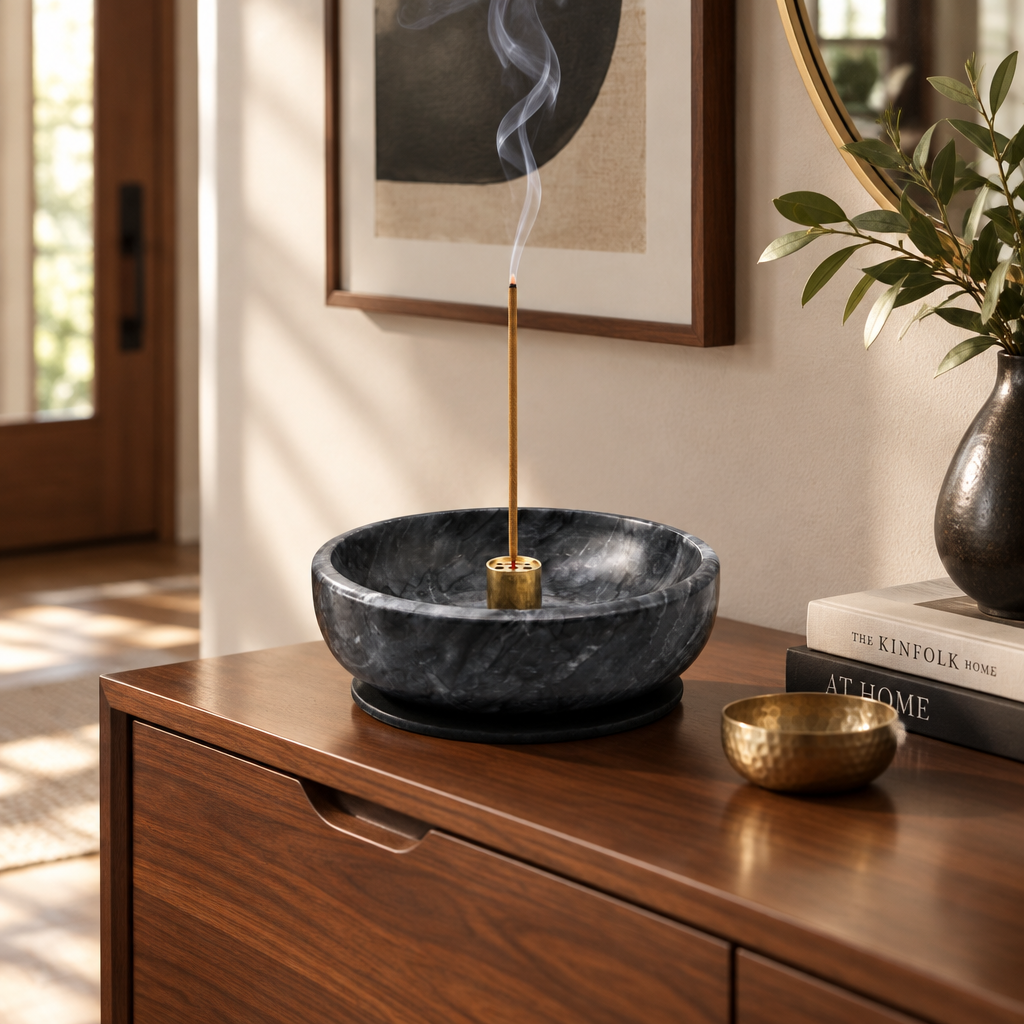 A dark charcoal stone incense burner bowl with a smoking incense stick, styled on a wooden console table next to a brass bowl and books in a warm, modern interior.